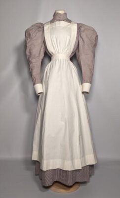 1890s style mauve and white striped dress on a mannequin, with very big mutton leg sleeves and a very small waist. The dress has a white apron over the top.